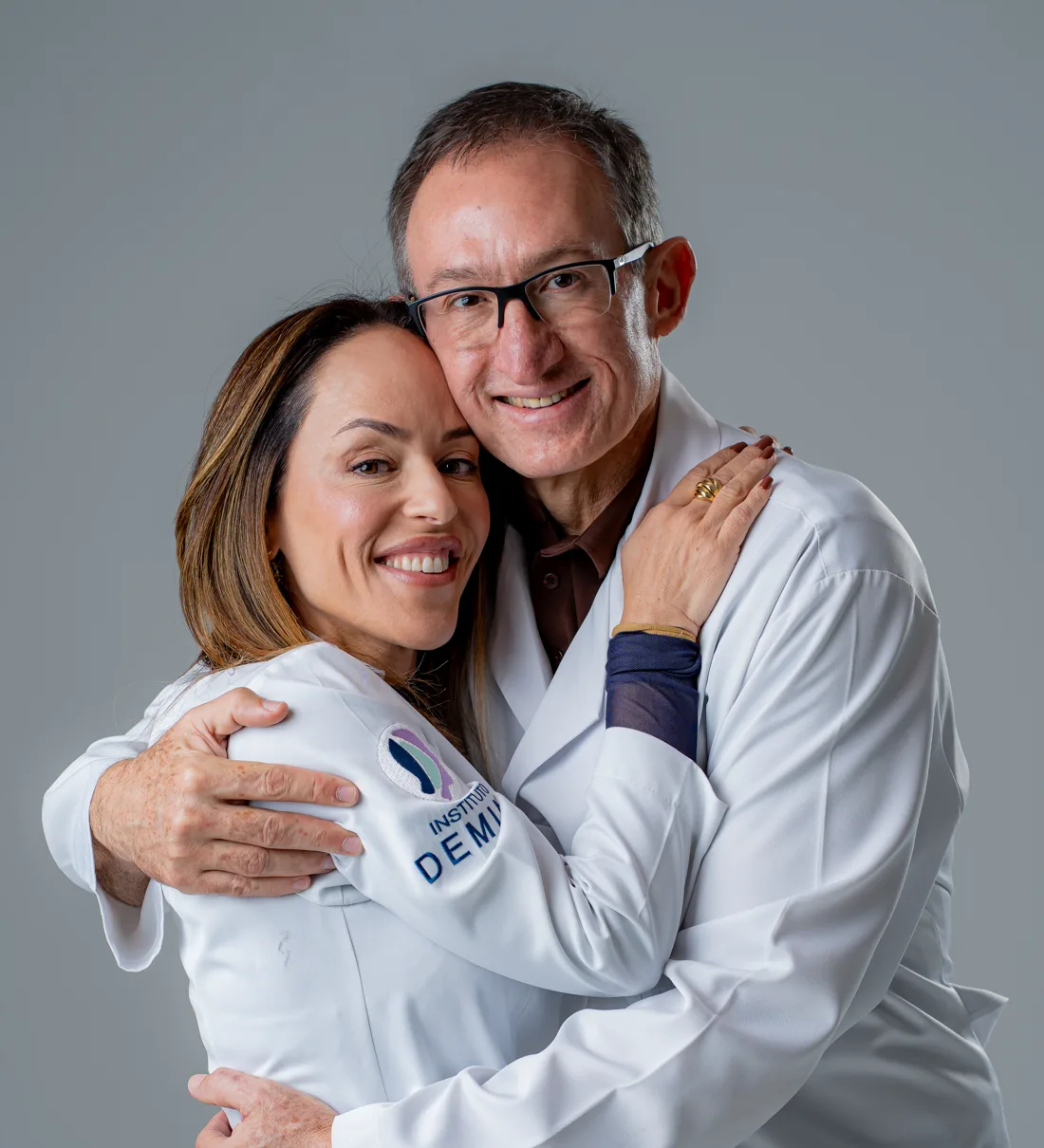 Dra. Mônica Demian e Dr. Marcelo Demian, de jaleco, abraçados e sorrindo